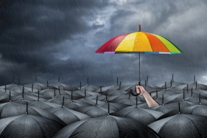 One rainbow coloured umbrella being held above lots of black umbrellas