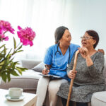 Health visitor and a senior woman during home visit. A nurse or a doctor examining a woman. Senior Woman Sitting In Chair  With Nurse In Retirement Home