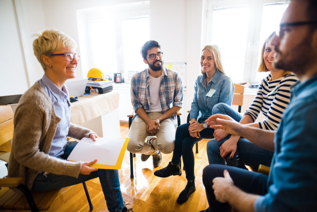 Group of diverse adults sat on chairs in a semi circle talking with each other. 