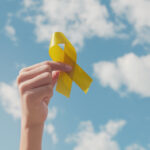 Hands holding yellow gold ribbon over blue sky, Sarcoma Awareness, Bone cancer, childhood cancer awareness, September yellow, World Suicide Prevention Day, endometriosis day concept