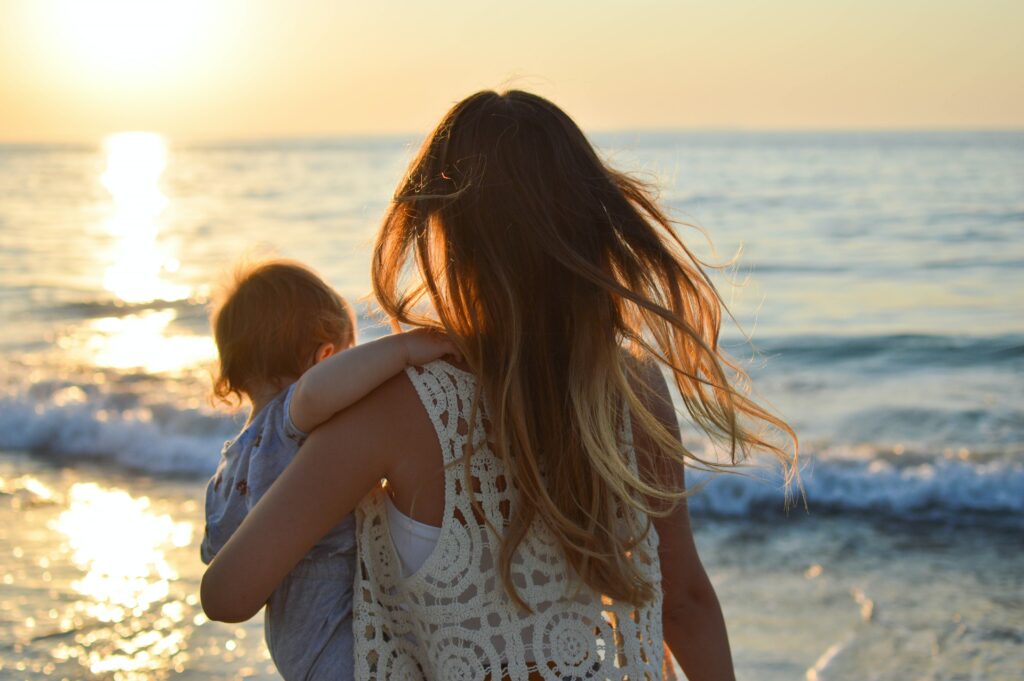 a woman with long hair and a white top faces a sunset on the beach while hold her baby in her arms, both are facing away