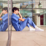 Tired,Young,Male,Doctor,Wearing,Scrubs,Sitting,Against,Wall,In