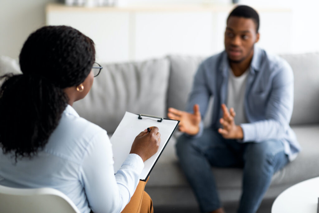 Two people are talking, one sat on a sofa gesticulating with his hands and the other is a woman holding a clipboard. Both are black
