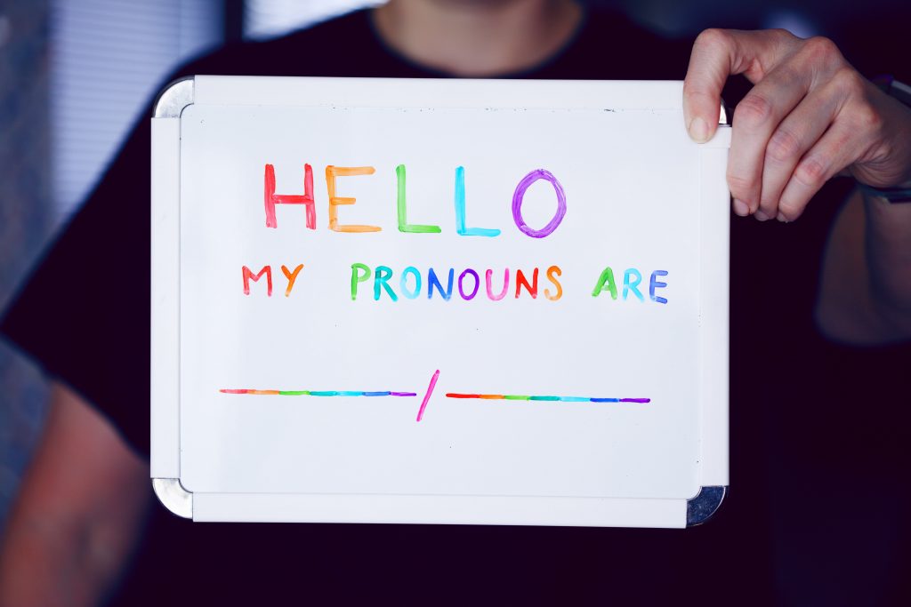 A whiteboard held up with the writing 'Hello, my pronouns are blank slash blank' in rainbow coloured writing