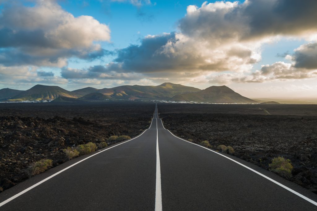 A long road heads off into the horizon, where there are cloudy blue skies and mountains