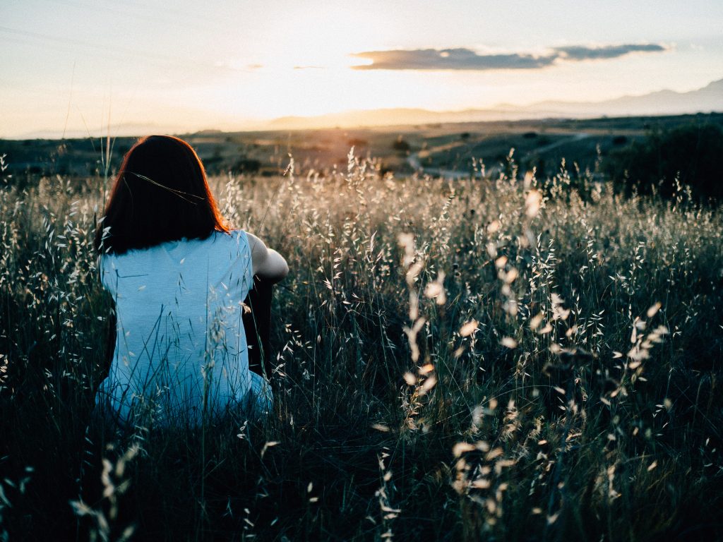 a girl with brunette hair crouches down in long grass watching a sunset