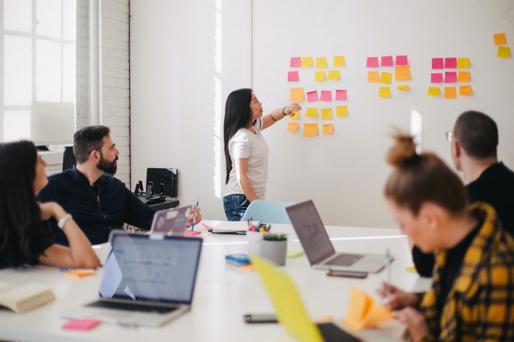  group sat around a large white table, someone is at the front of the room presenting to the group and pointing to several pink and orange post it notes stuck on the wall. 