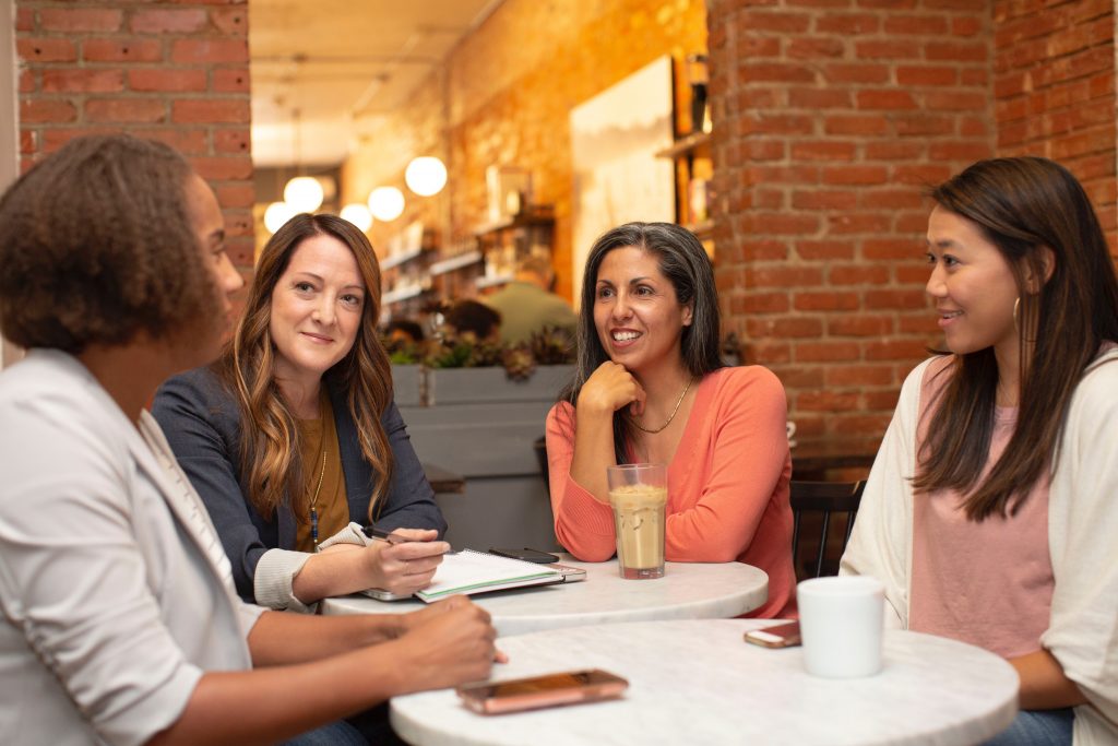 four young professionals sat down around a desk with coffees and papers in front of them, smiling. 