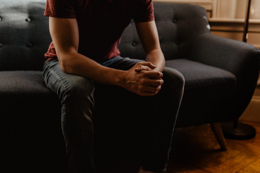 a muscular man in a red t shirt and jeans sits on a sofa, learning forward, his face out of view, as if speaking to someone opposite