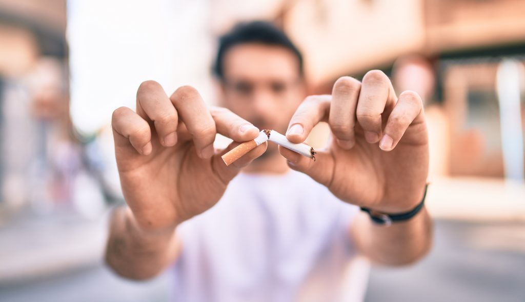 a man in the back ground is blurred but holds his hands out in front of him breaking a cigarette in half in the foreground
