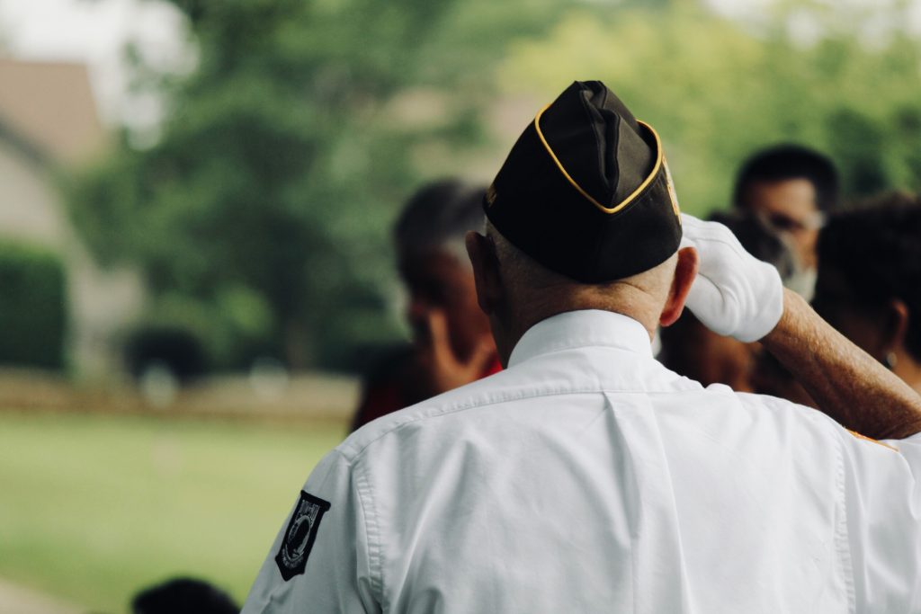 an older man faces away from the camera, wearing a military beret, a crispy white shirt with a black badge on the arm, with his hand in a salute