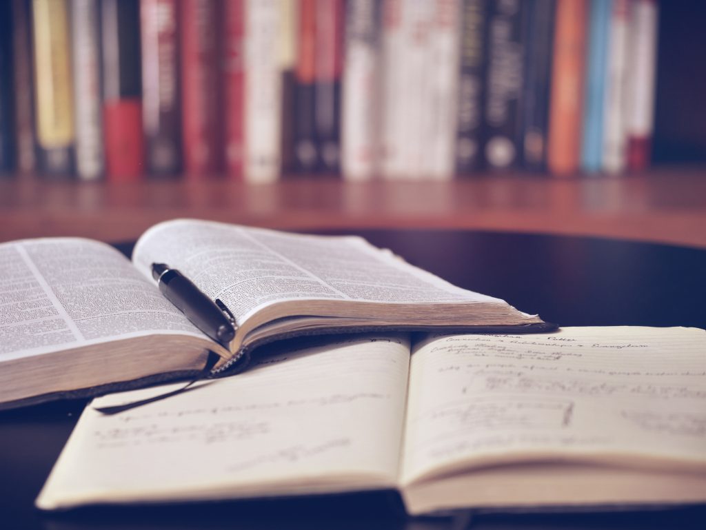 Two books lie open on a table, one has printed text on it, the other is a notebook with hand written notes. 