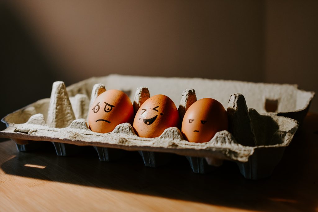 three eggs in a box have three different emotions drawn on them in black pen, sad or scared, laughing and annoyed