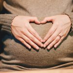 a pregnant woman in a grey top makes a heart shape with her hands over her pregnancy bump