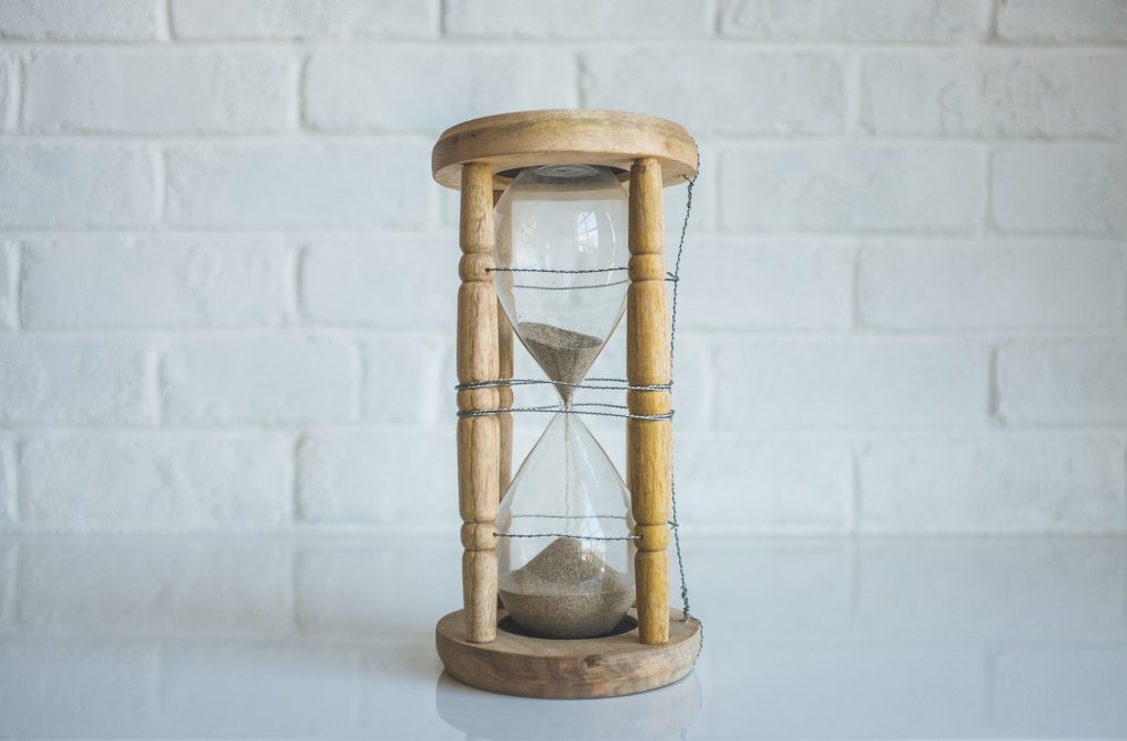 a sand timer with off white coloured sand is turned upside down, on a white table in front of a white brick wall