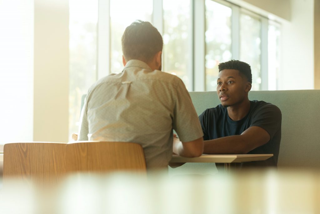 two men sit opposite each other at a table, one in a shirt and one in a black tshirt, the man with his back to us is white with brunette hair and the man facing us is Black with short dark hair. 