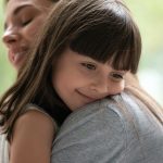 Side view close up head shot happy little adopted kid girl put head on mothers shoulder, feeling love and support. Small cute daughter hugging embracing cuddling young smiling mother at new home.