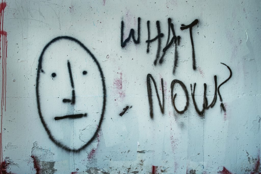 graffiti in black paint reads 'what now' in capitals next to a cartoon face