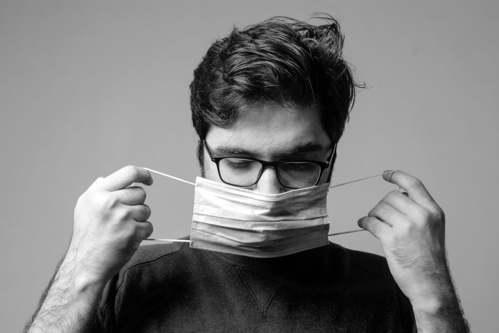 black and white photo of a man with dark hair and glasses putting on a surgical mask