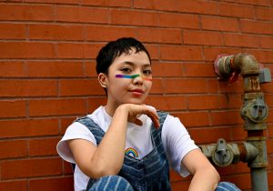 Young person sat against wall with pride face paint.