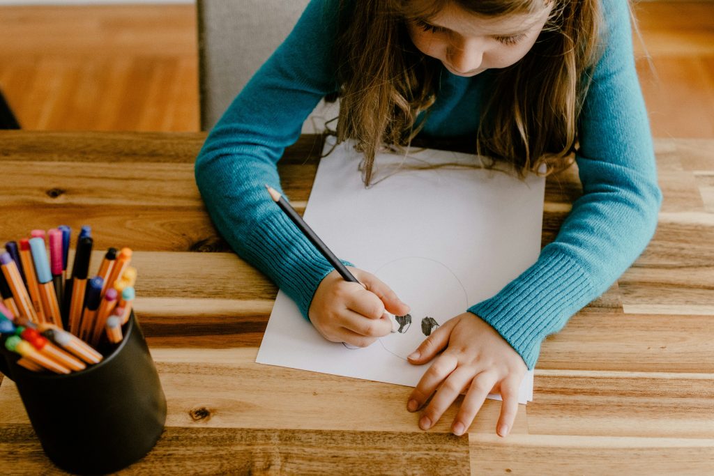 a girl in a blue long sleeve top with brown hair is drawing on a white sheet of paper with a pencil
