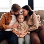 Daughter,With,Neutral,Face,Sits,Hugged,By,Parents,,Kissed,Gently,