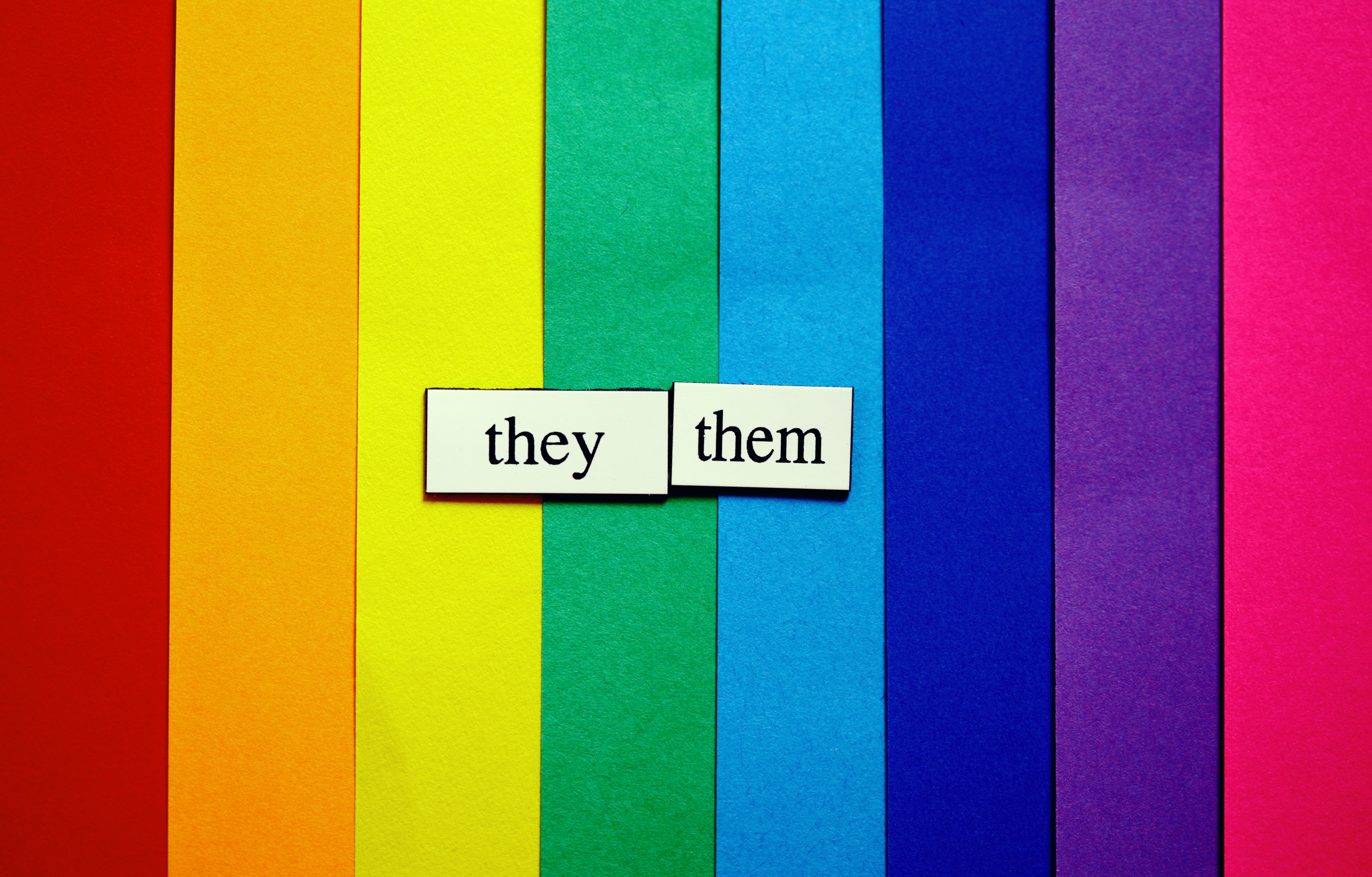 a multi coloured rainbow with two white stickers in the centre that say 'they' and 'them'