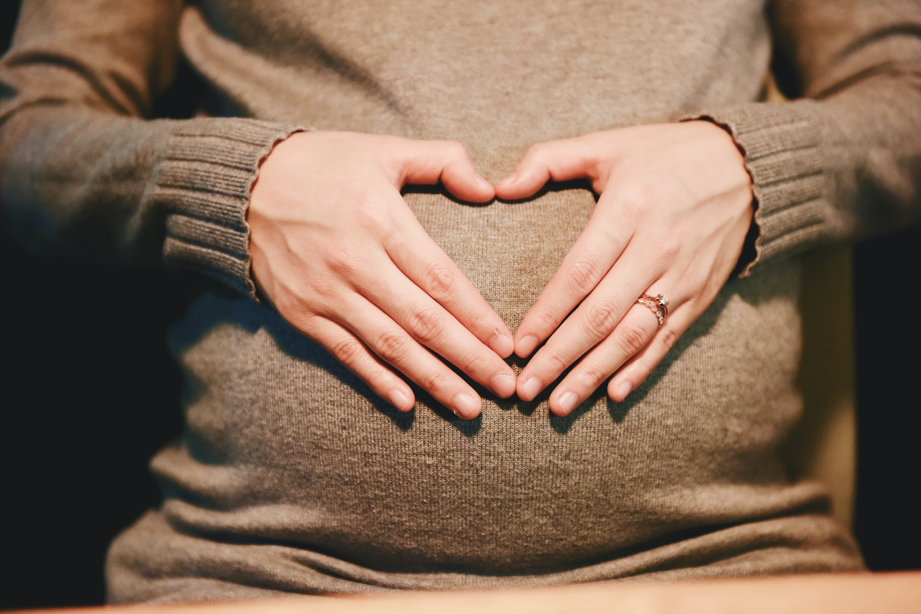 a pregnant woman in a grey top makes a heart shape with her hands over her pregnancy bump